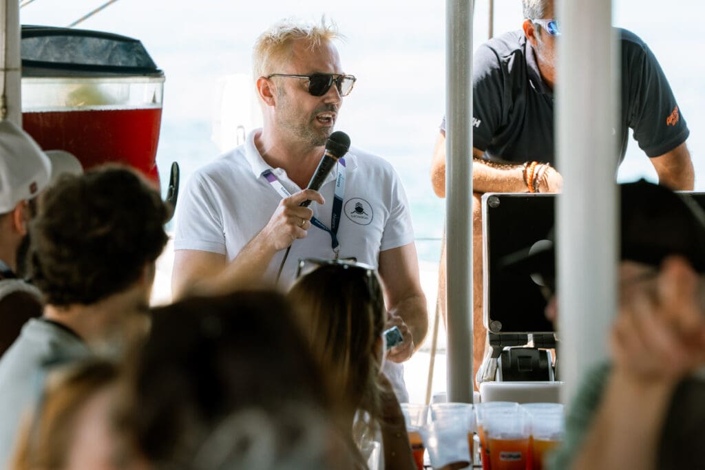 The host speaking into a microphone to guests seated across the deck.