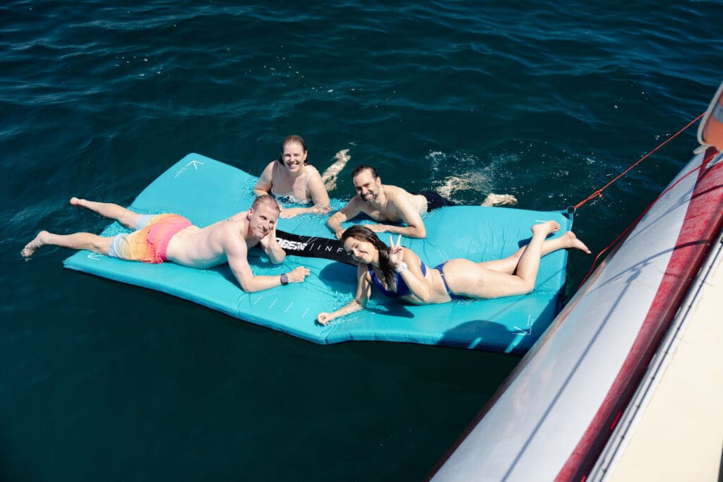 Guests sitting on a turquoise floating mat beside the hull of the catamaran.